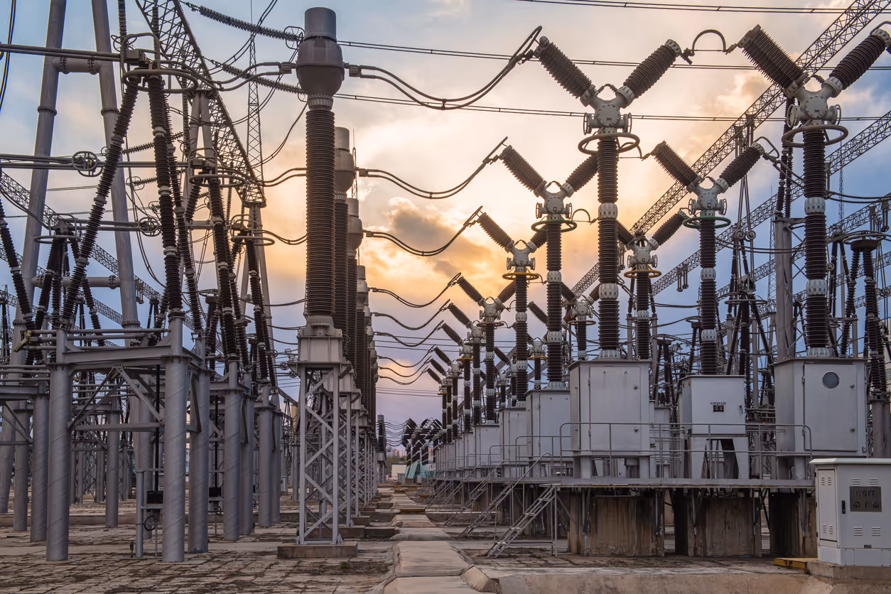 Rows of electrical transformers and insulators at a power substation under a partly cloudy sunset sky.