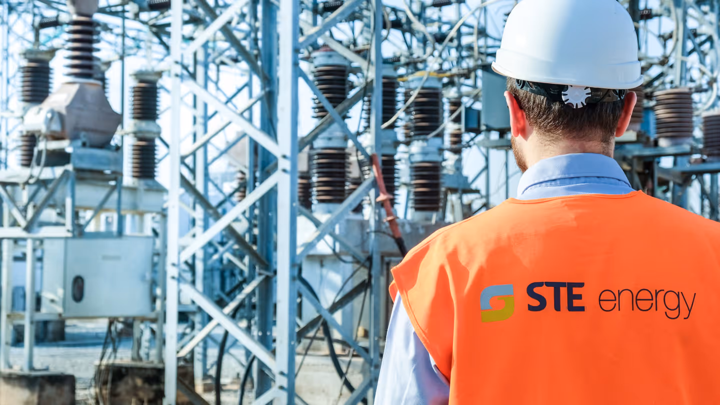 Worker wearing an orange vest and white hard hat inspecting electrical substation equipment.