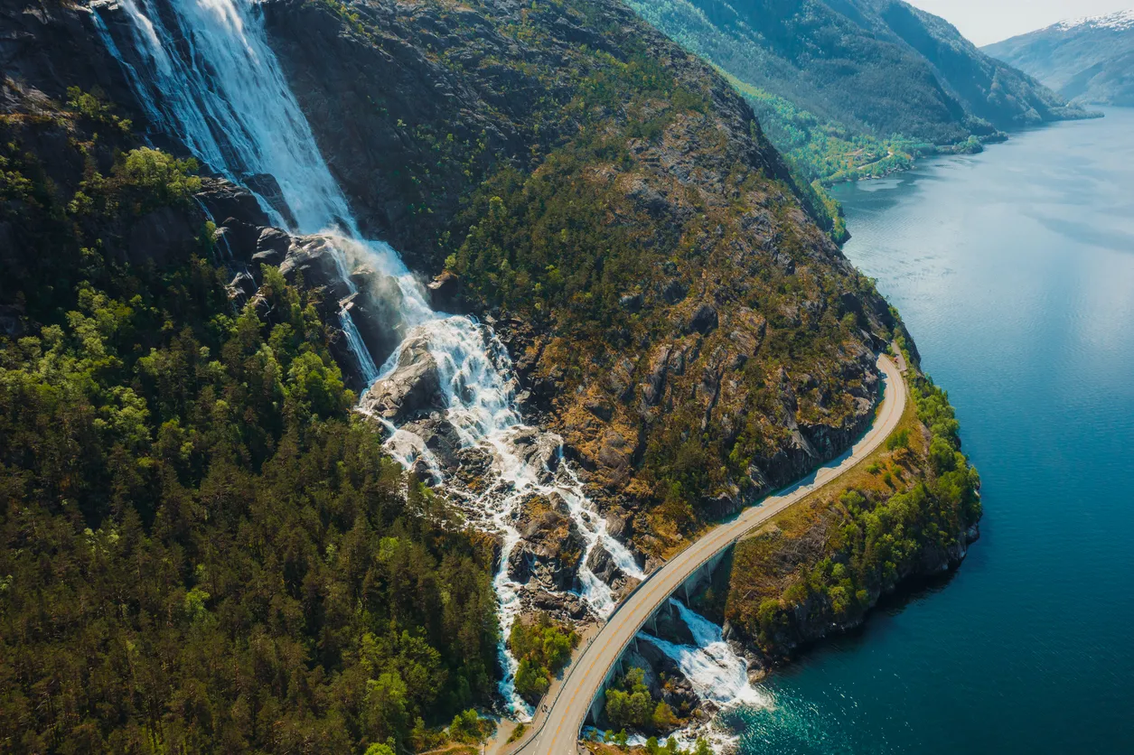 Curved road crossing over a waterfall cascading down rocky cliffs into a lake surrounded by forested mountains.
