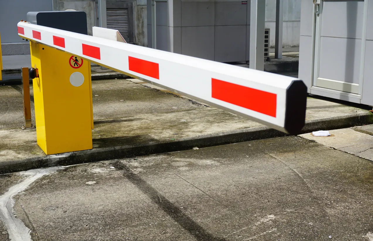 White and red striped automatic vehicle barrier arm attached to a yellow base in an outdoor parking or entrance area.