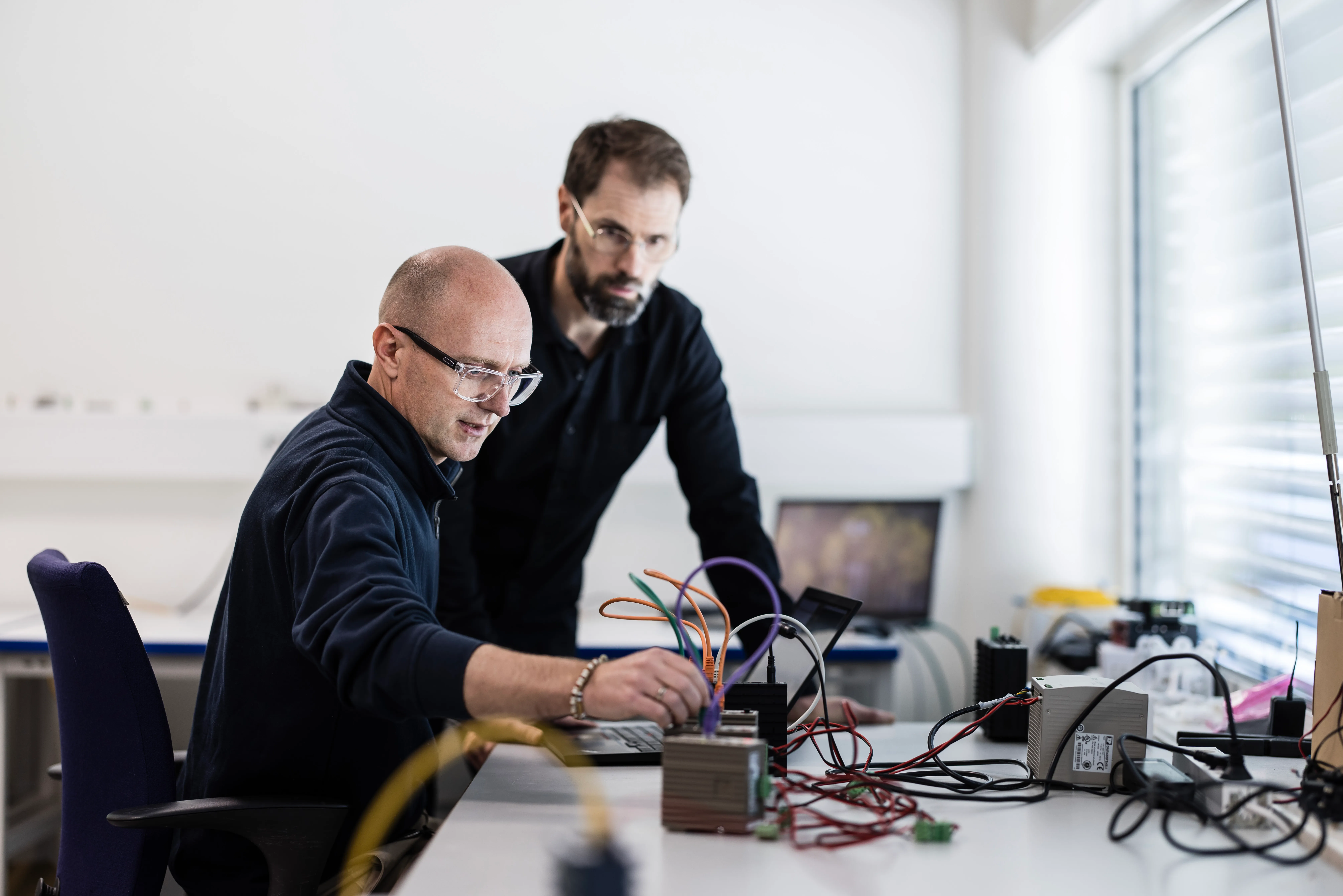 Two men working with electronic equipment and cables on a desk in a well-lit office.