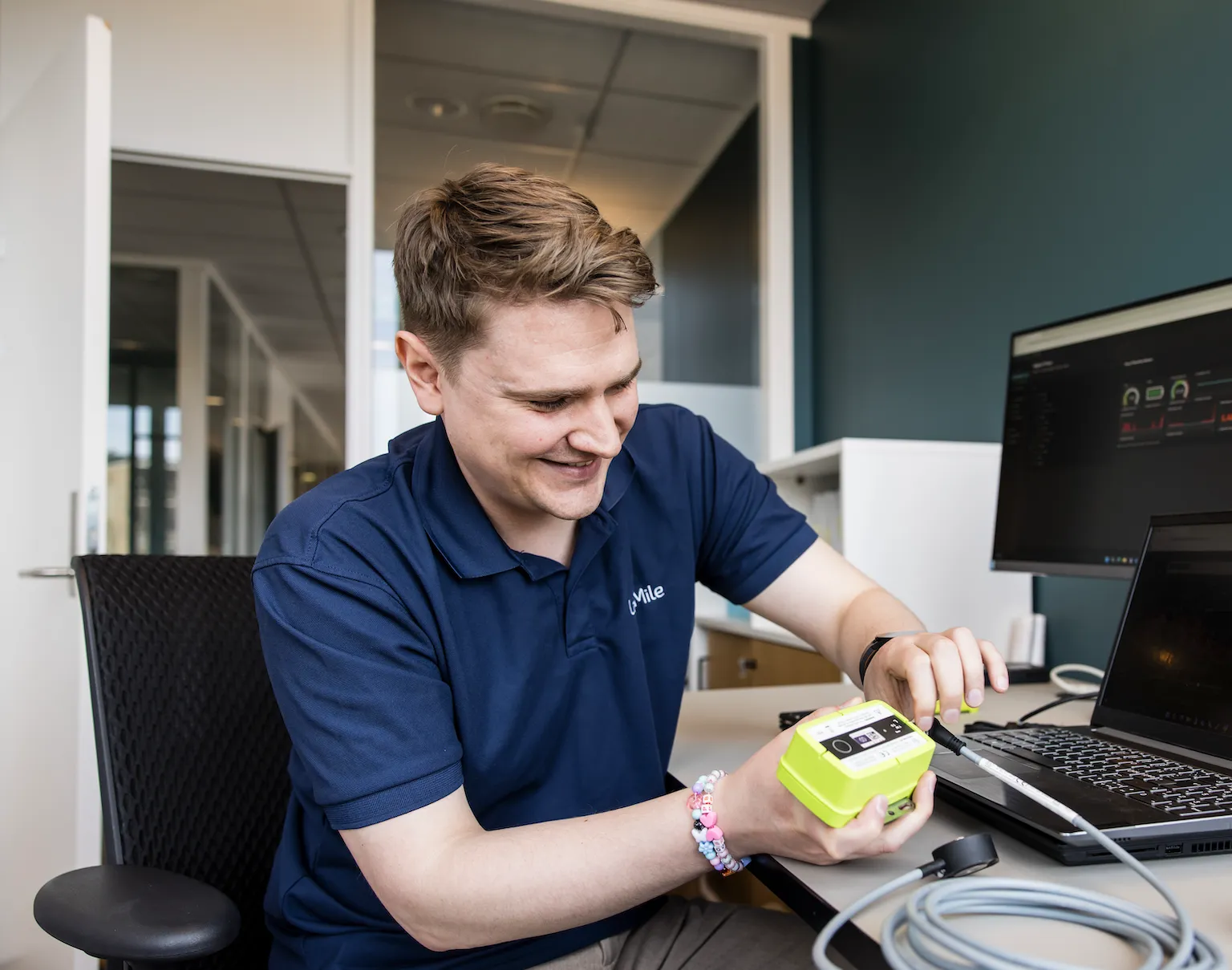 Man in blue polo shirt working with a small yellow electronic device connected to a laptop in an office setting.