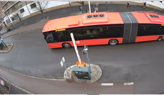Red articulated bus driving on a city street with a traffic barrier and traffic signs in the foreground.