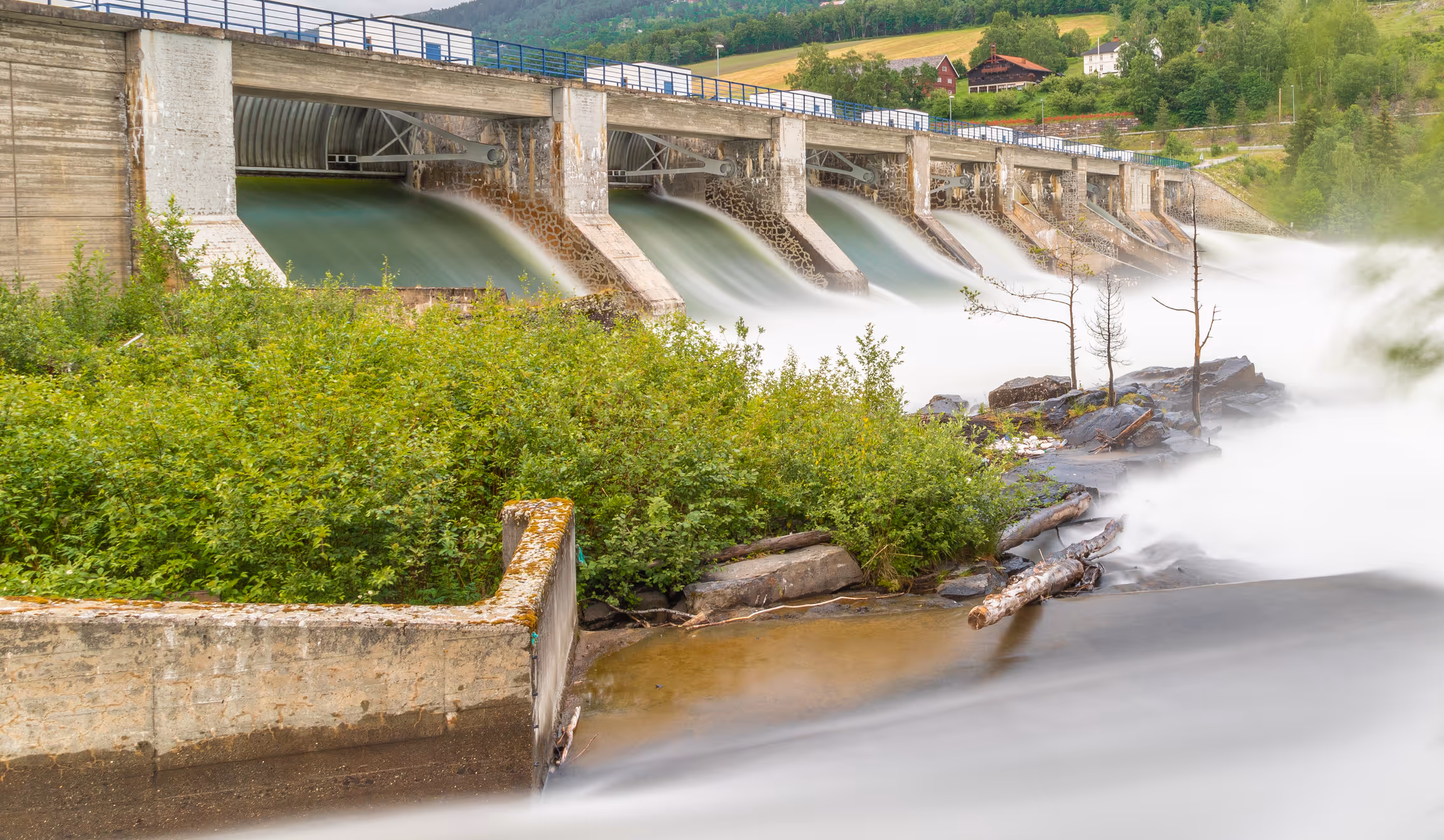 Concrete hydroelectric dam releasing flowing water with green vegetation and rocky riverbank in a rural landscape.