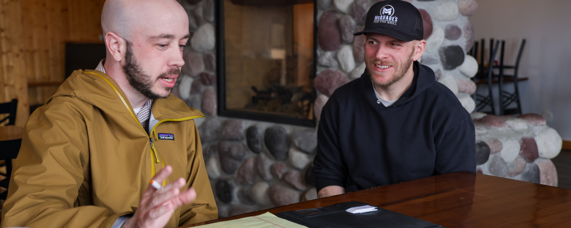 Two people seated at a wooden table in front of a stone fireplace, with a folder and placemat on the table.