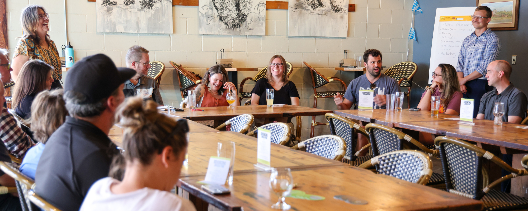 People seated around large wooden tables in a meeting space with artwork on walls and a whiteboard in the background.