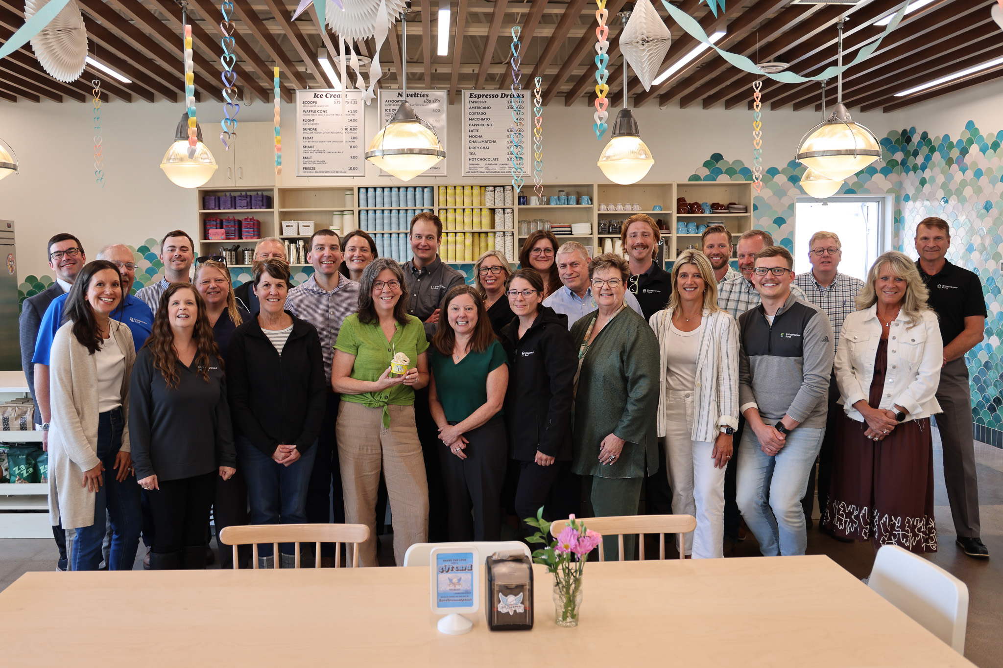 A large group photo of the EFund team at Love Creamery's Superior location. The team is casually dressed, while enjoying ice cream with the ice cream counter in the background. There are multiple rows of people: the front row is crouching or kneeling, and the back row is standing. Everyone is smiling and looking at the camera, indicating a friendly and cohesive team atmosphere.