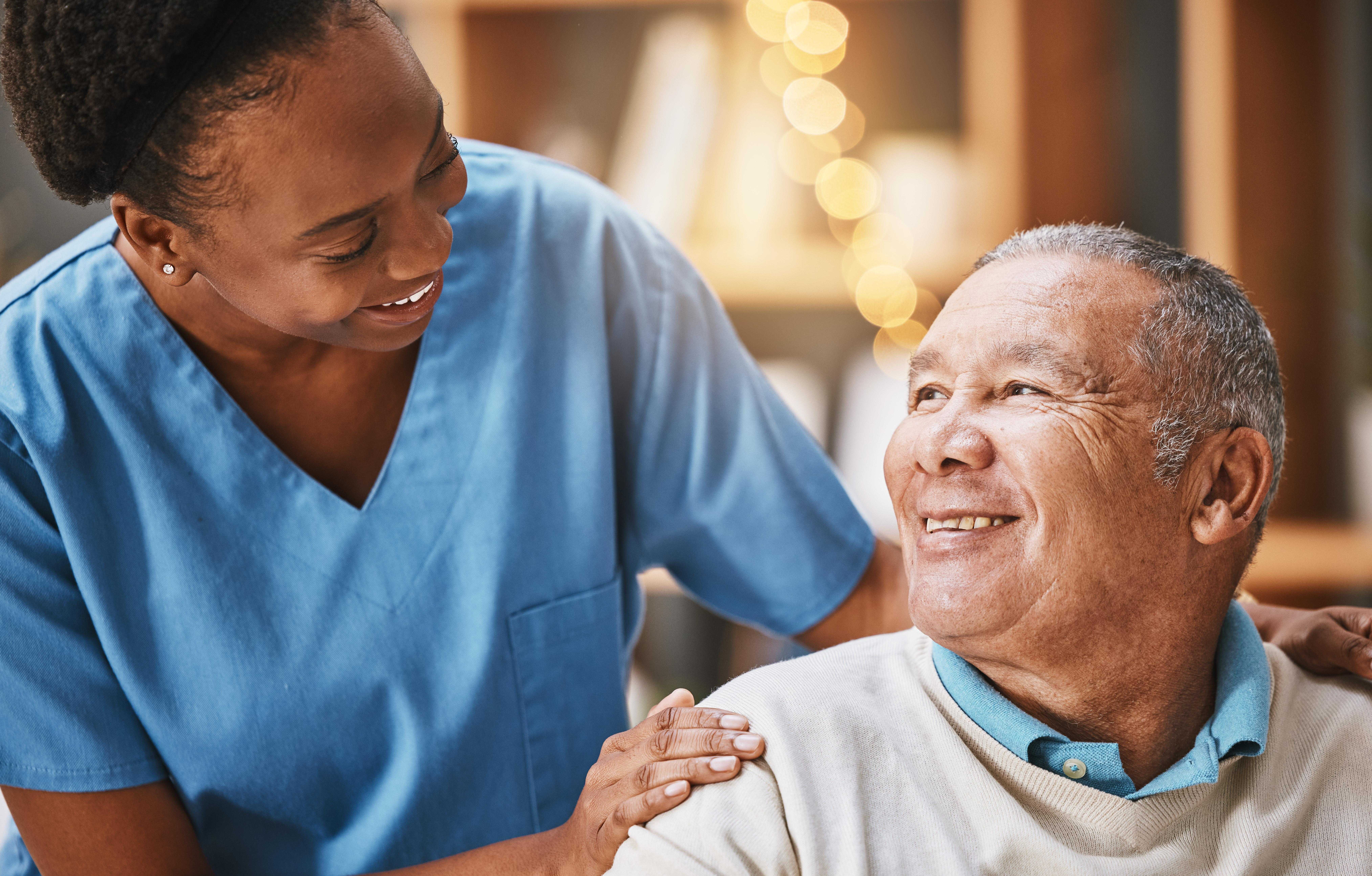 Smiling nurse in blue scrubs comforting an elderly man who is looking up at her with a smile.