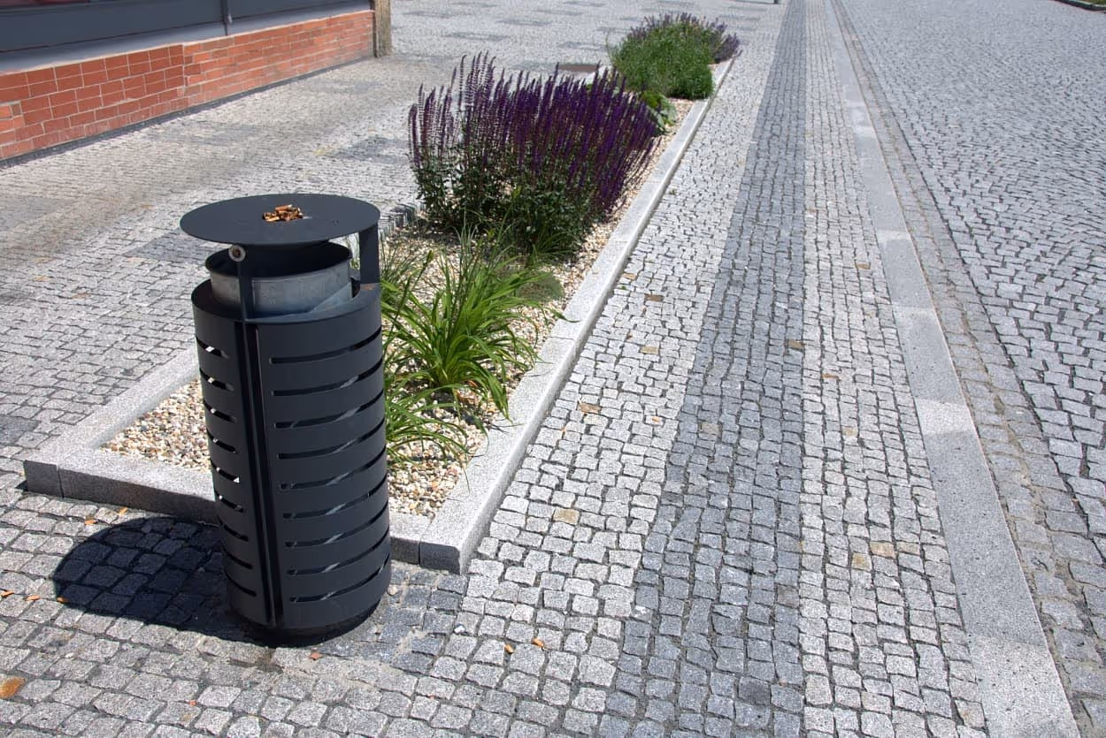 Žulová štípaná dlažba s odvodňovacím pásem, žulovými obrubníky a zeleným ostrůvkem s květinami, doplněná odpadkovým košem.