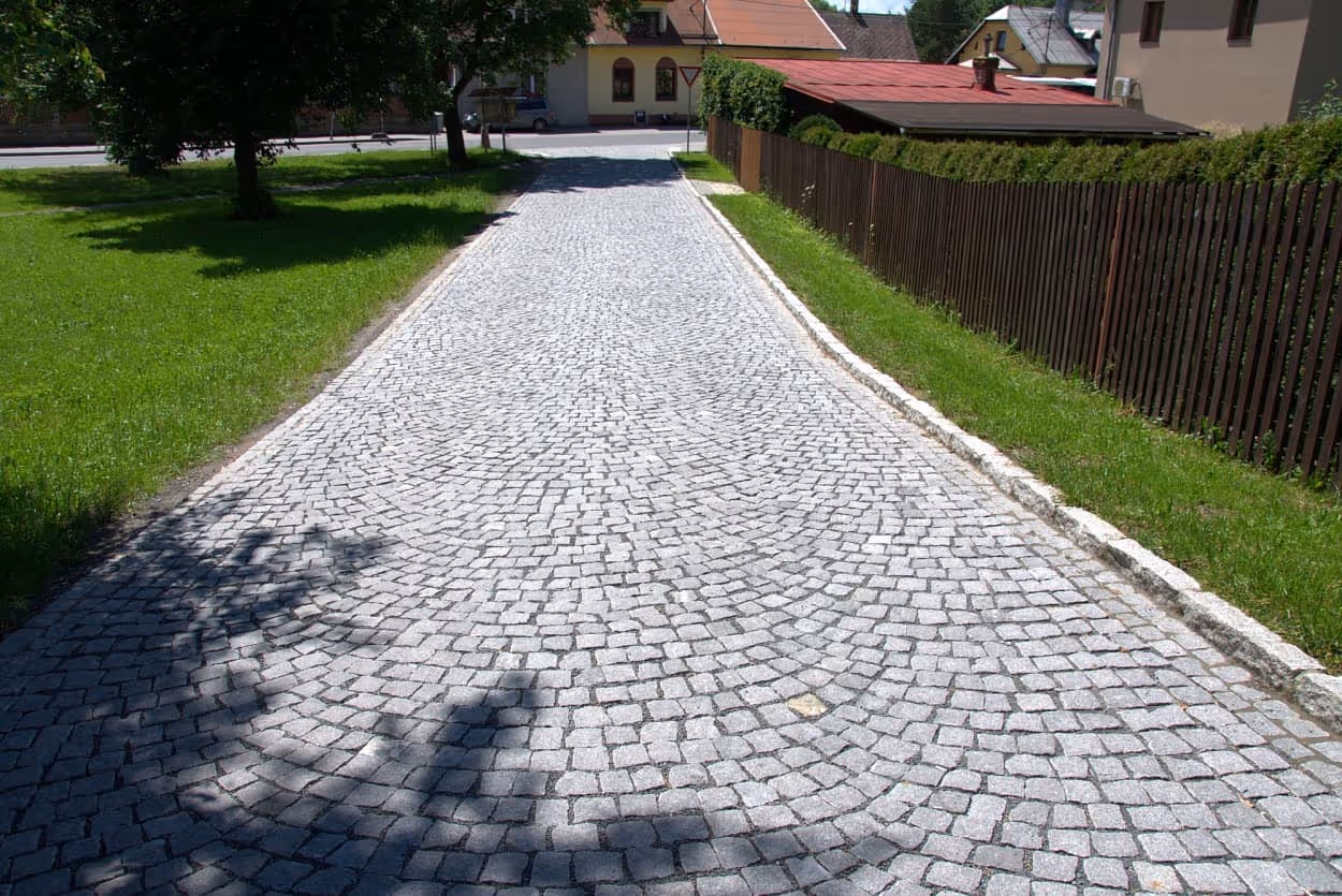 Cesta vydlážděná šedou štípanou žulovou kostkou v obloukovém vzoru, lemovaná žulovými obrubníky a obklopená zelení.