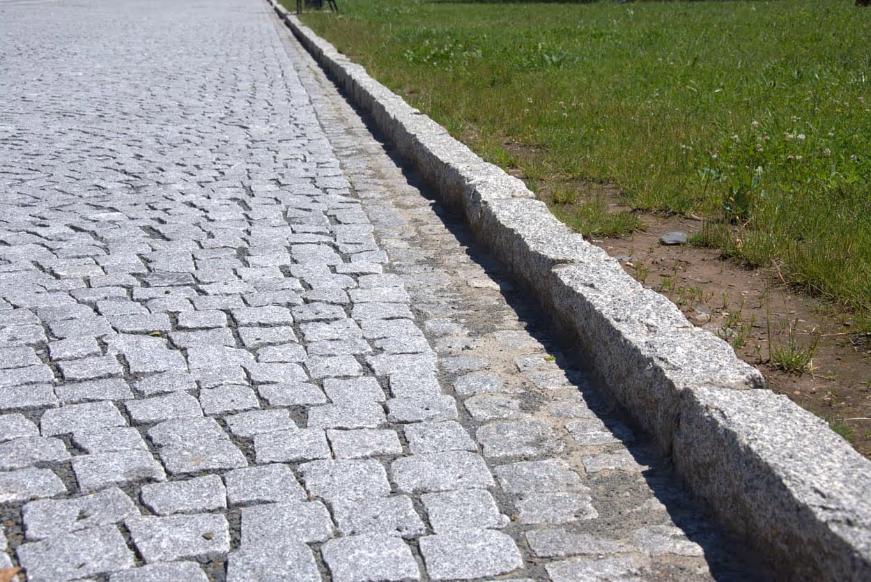 Detail žulové štípané dlažby s obrubníkem, který odděluje vozovku od travnaté plochy. Odolné řešení pro cesty.