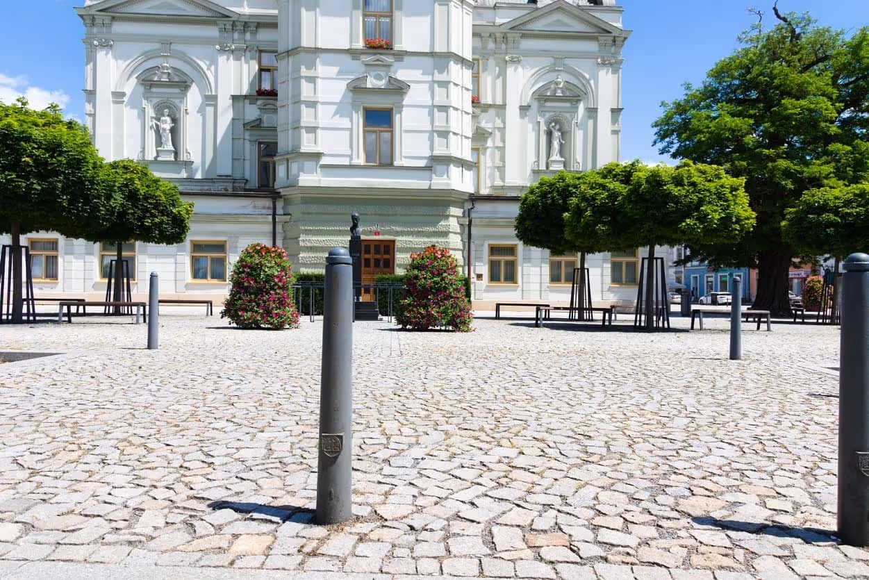 Náměstí před historickou budovou s vydlážděným povrchem z přírodního kamene a moderními sloupky.