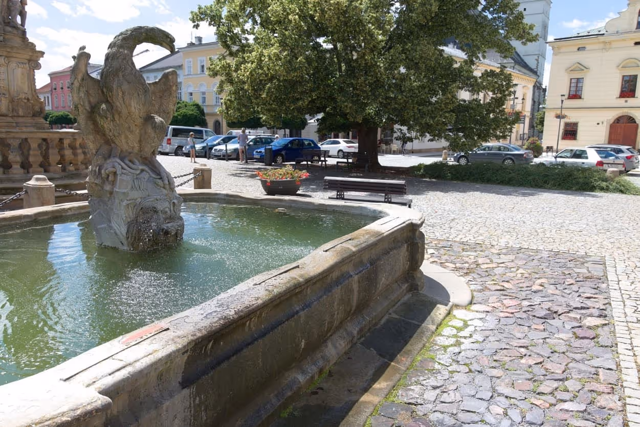 Kamenná kašna s vodní sochou, obklopená žulovou dlažbou a stromy v historickém centru Uničova.