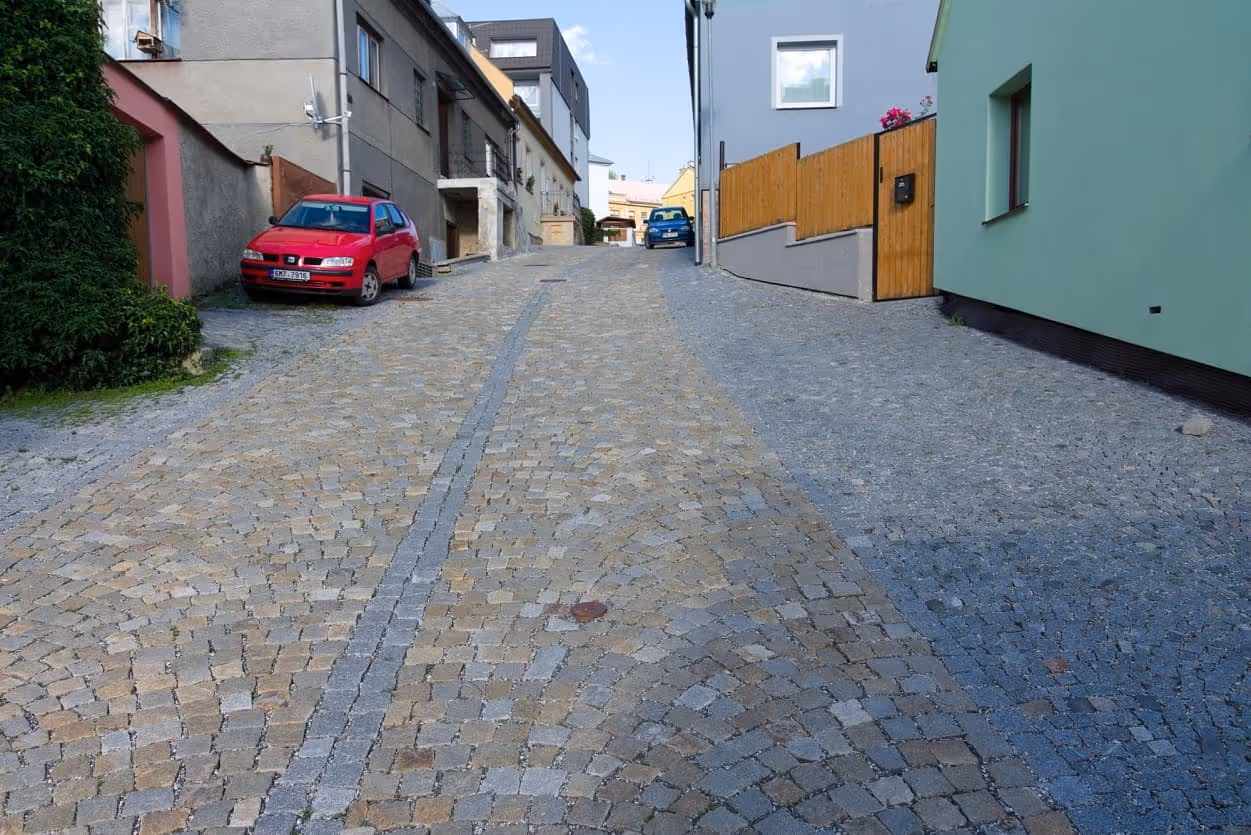 Úzká ulice dlážděná šedožlutými žulovými kostkami s centrálním odvodňovacím pásem, obklopená moderní i tradiční zástavbou.
