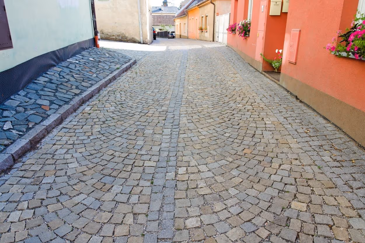 Dlážděná ulička s šedožlutými žulovými kostkami, lemovaná domy s květinami v oknech a zvýšeným chodníkem z lomového kamene.