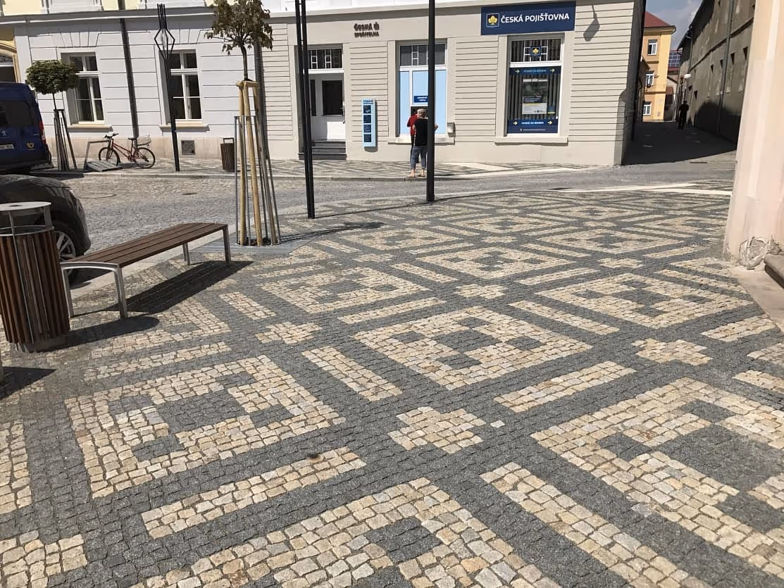 Ornamentální dlažba z žulových kostek na náměstí, doplněná lavičkami, odpadkovými koši a mladými stromy.