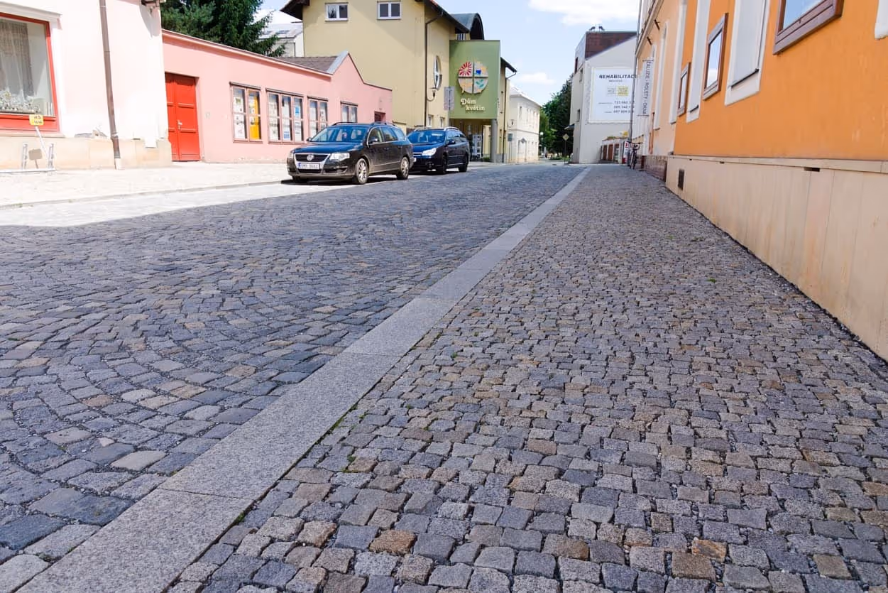 Ulice s dlažbou z žulových kostek, doplněná žulovým obrubníkem a parkovanými auty podél chodníku.