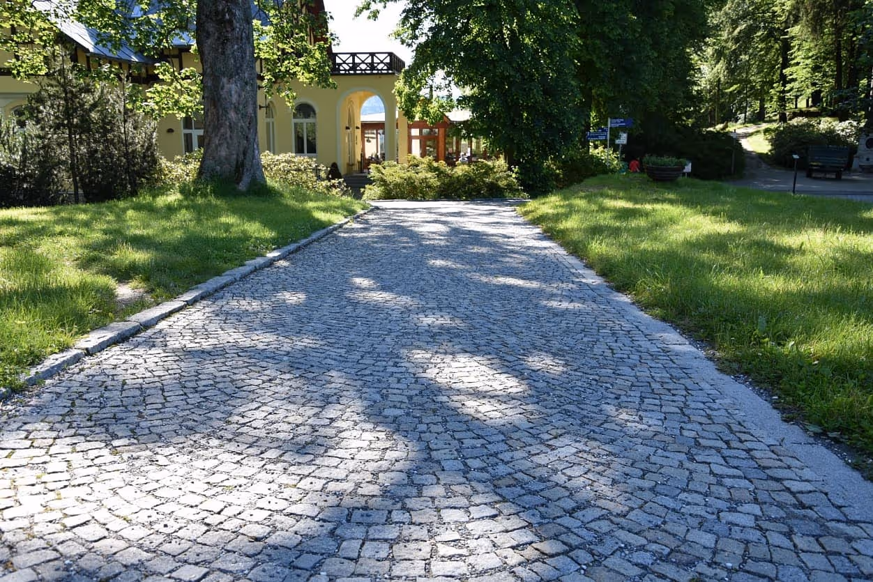Chodník z obloukově kladené žulové dlažby v Priessnitzových léčebných lázních, lemovaný zelení a historickou budovou.