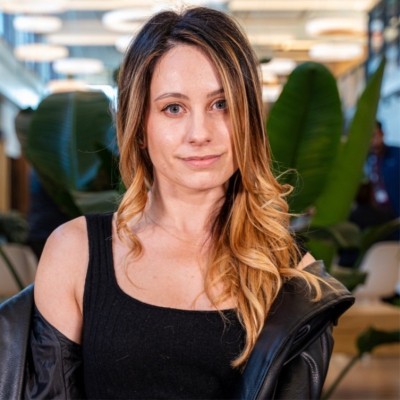 Young woman with long blonde hair wearing a black top and leather jacket, sitting indoors with green plants in the background.