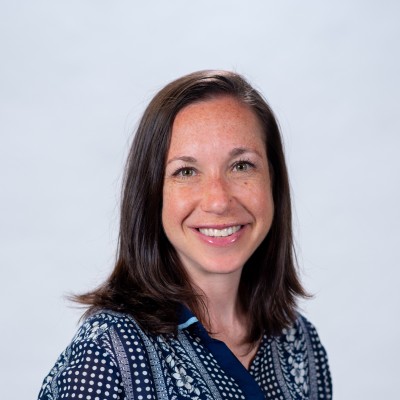 Smiling woman with shoulder-length brown hair wearing a patterned navy blue top against a light background.