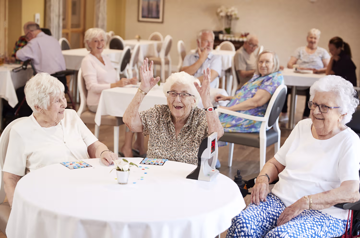 Elderly women sitting around a table playing bingo, one woman raising her hands joyfully while others smile.