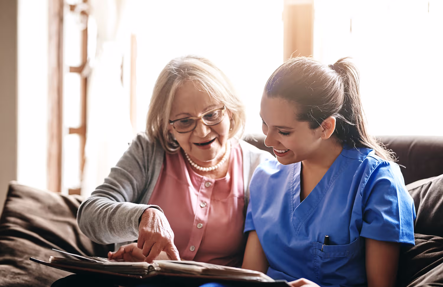 Elderly woman and healthcare worker in blue scrubs smiling while looking at a photo album together.