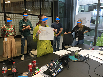 A photo of a woman showing to the camera the persona's artefacts she developed on a piece of paper. While people are behind her.