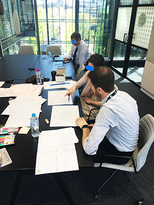 A photo of two people developing paper prototypes on a desk while another person observes and takes notes.