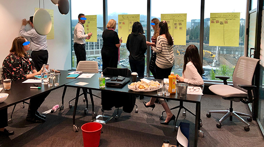 A photo of a group of people discussing and placing sticky notes on Post-it Easels placed on a window and a wall.