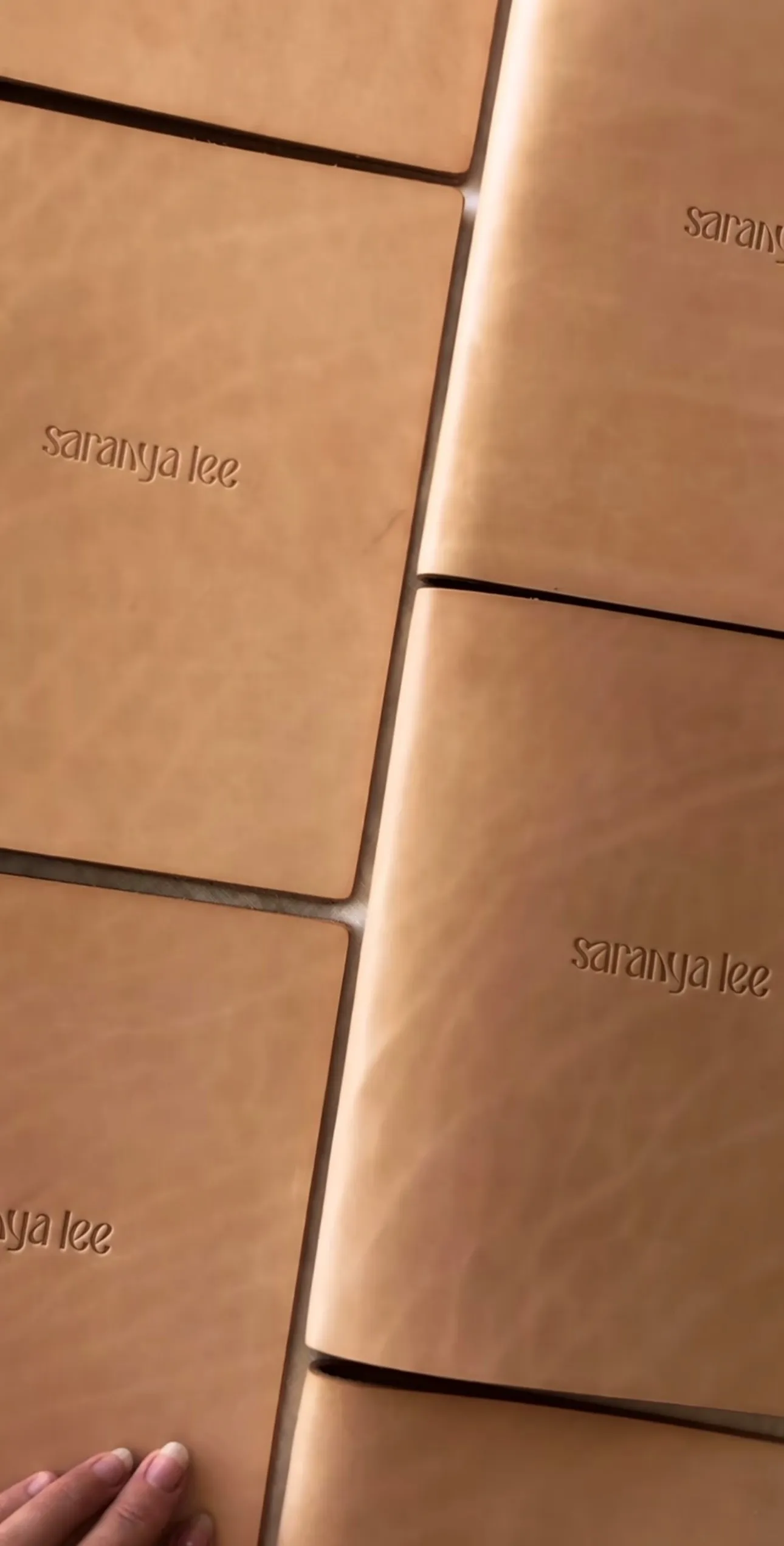 Several brown leather-bound menus embossed with 'saranya lee' stacked on a surface, with a hand touching one menu.
