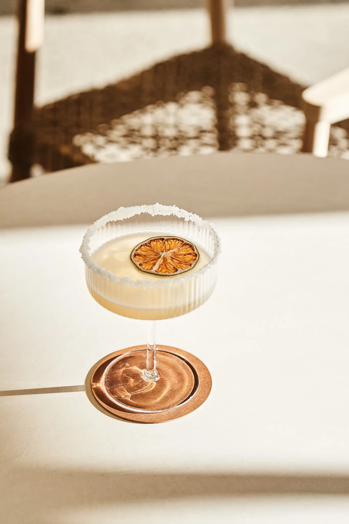 Cocktail glass with salted rim and dried orange slice garnish on a light surface casting a circular shadow.