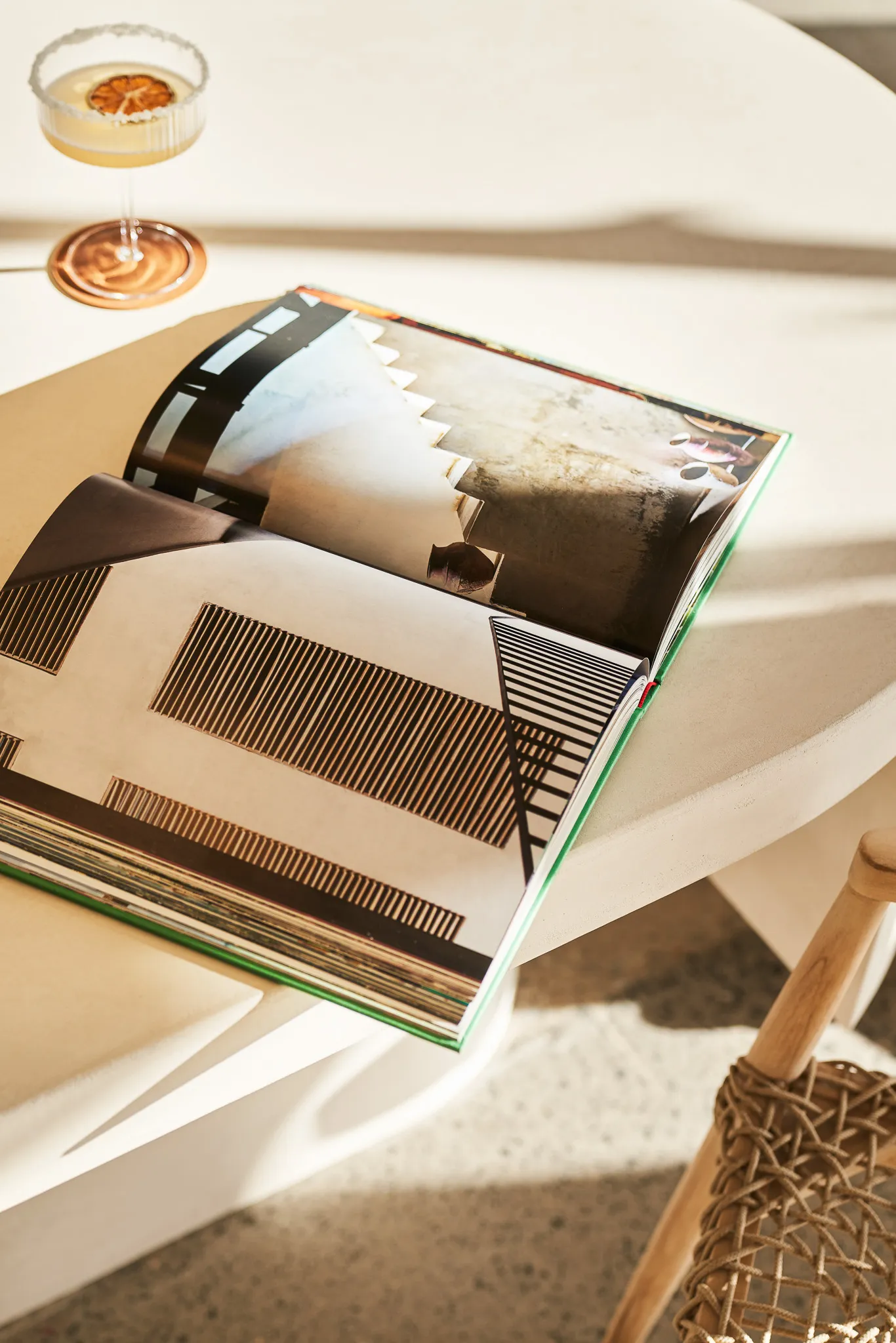 Open book with architectural photos on a round white table next to a cocktail glass and a woven chair.