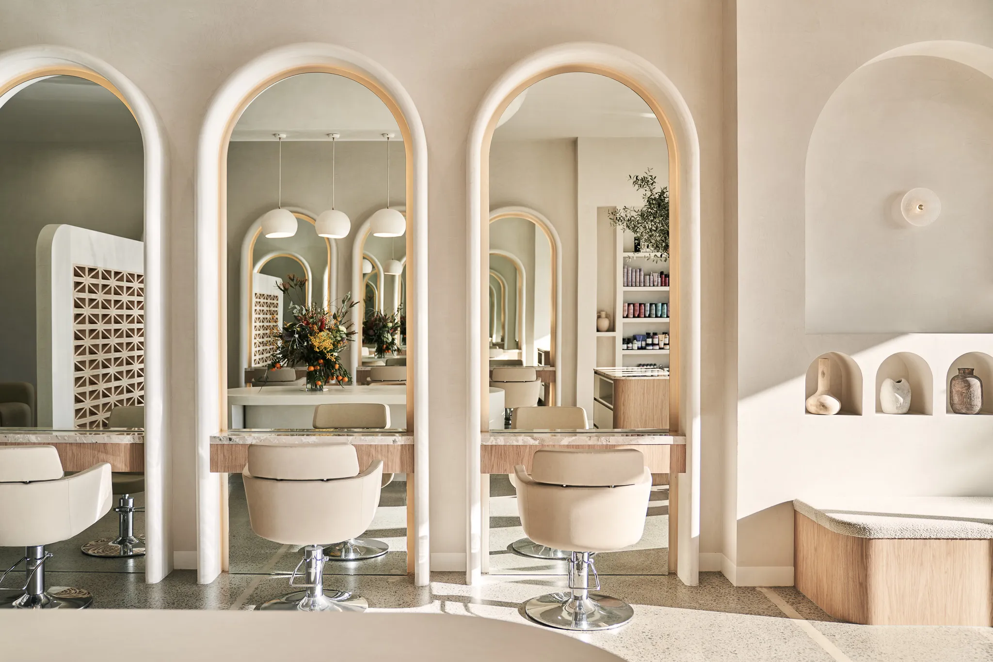 Bright, modern hair salon interior with beige chairs in front of large arched mirrors and a floral arrangement on the counter.
