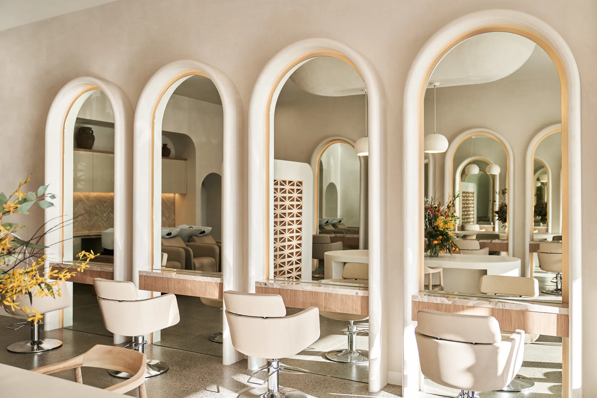 Bright modern salon interior with arched mirrors, beige styling chairs, and minimal decor including a floral arrangement.