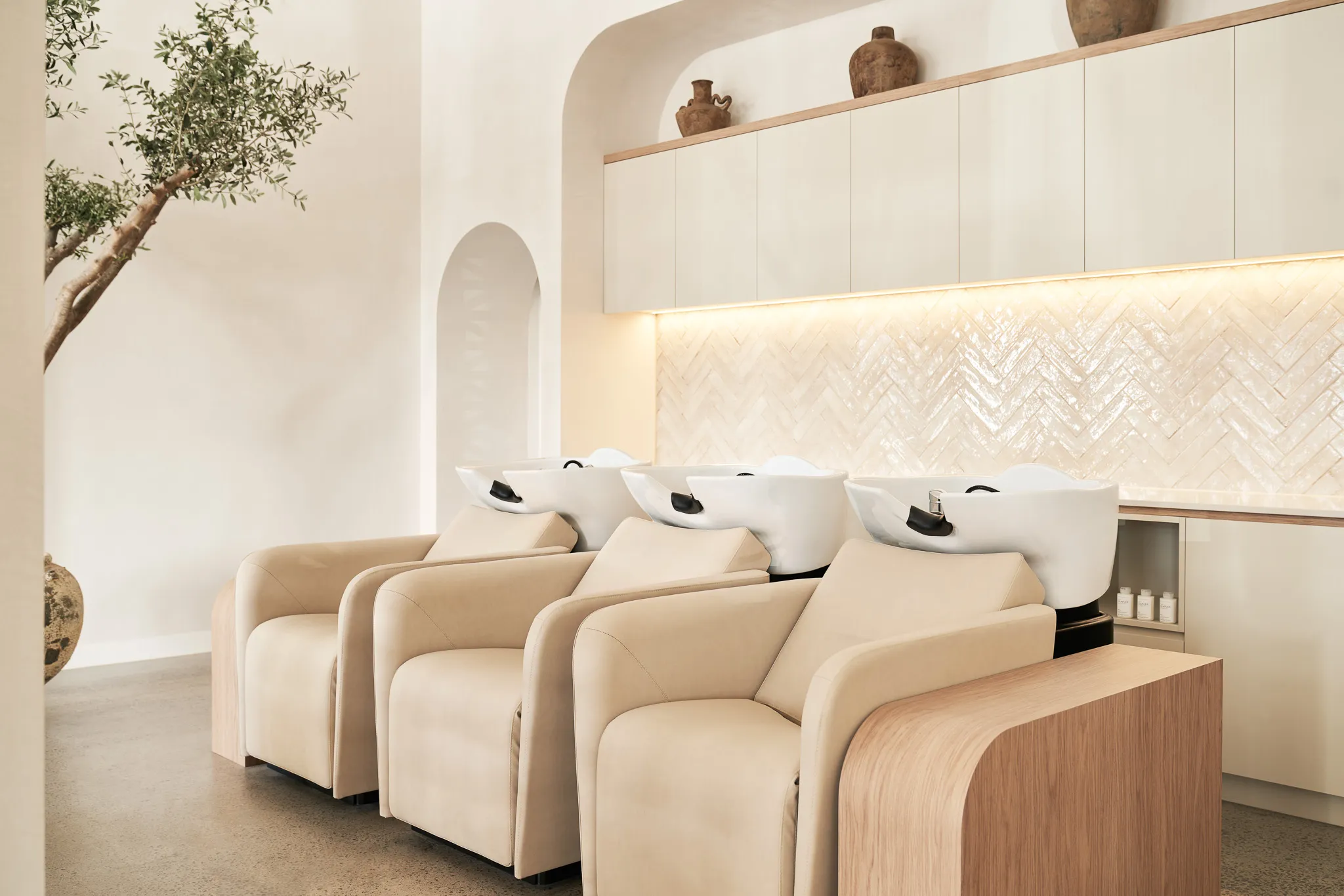 Minimalist salon interior with three beige leather shampoo chairs and white sinks against a tiled wall with soft lighting.