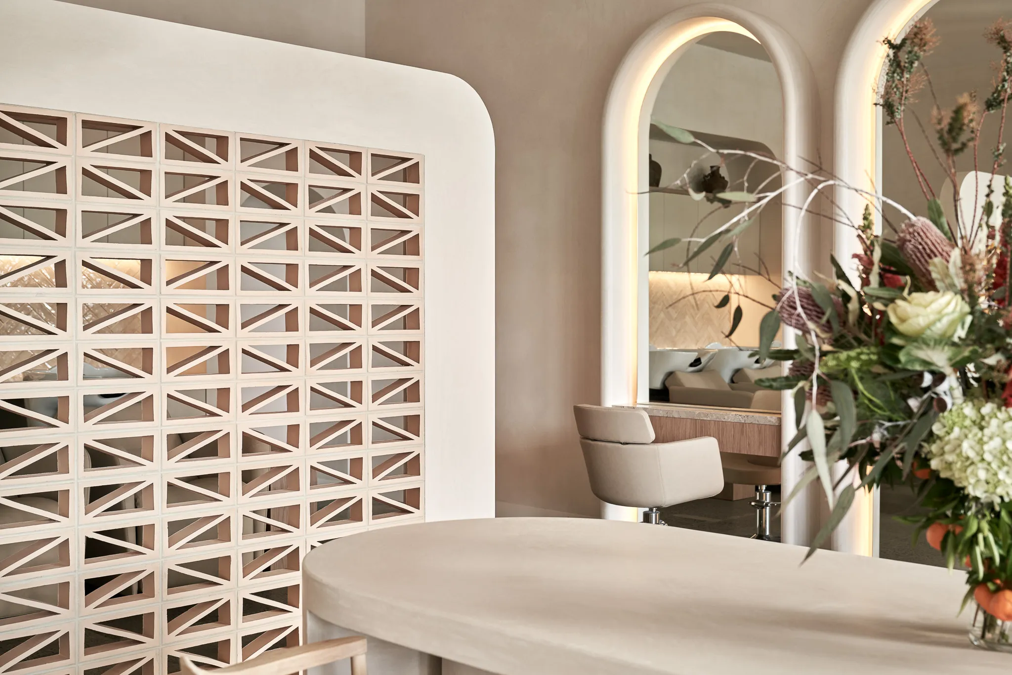 Modern interior with beige rounded table, decorative partition wall, illuminated arched mirrors, and floral arrangement.