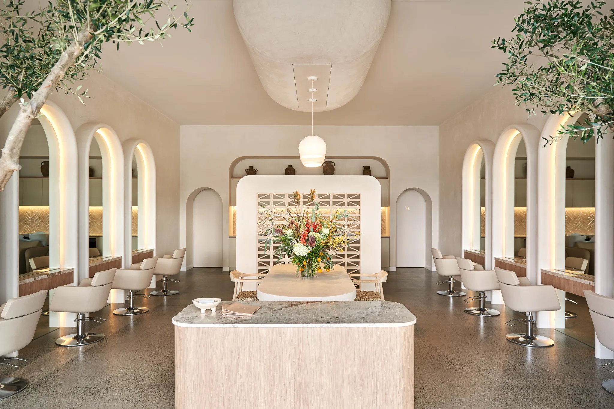 Modern salon interior with beige styling chairs, arched mirrors with backlighting, and a floral centerpiece on a central table.