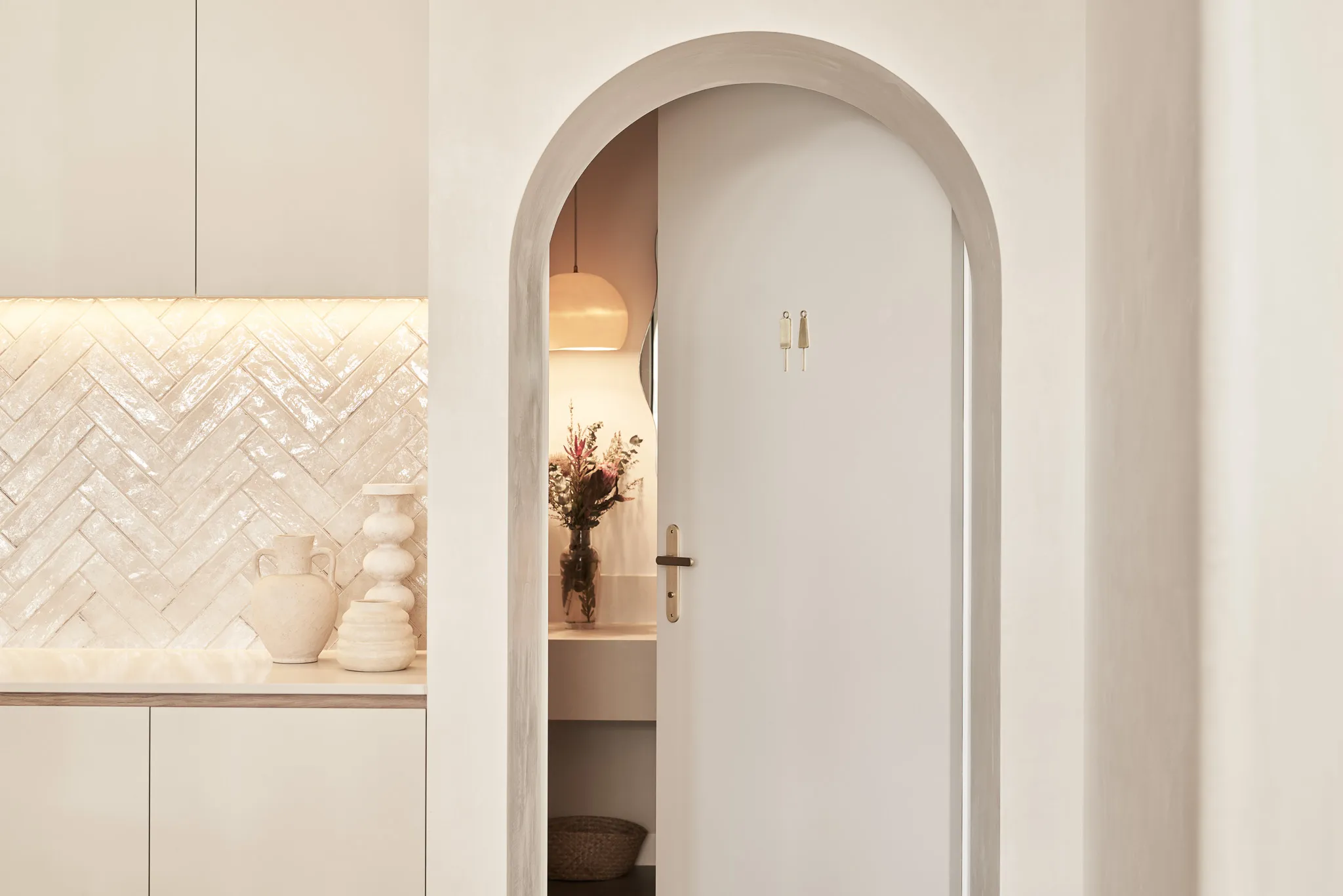 Light beige kitchen with an arched doorway partially open to a room featuring a vase of flowers on a shelf and a warm hanging lamp.