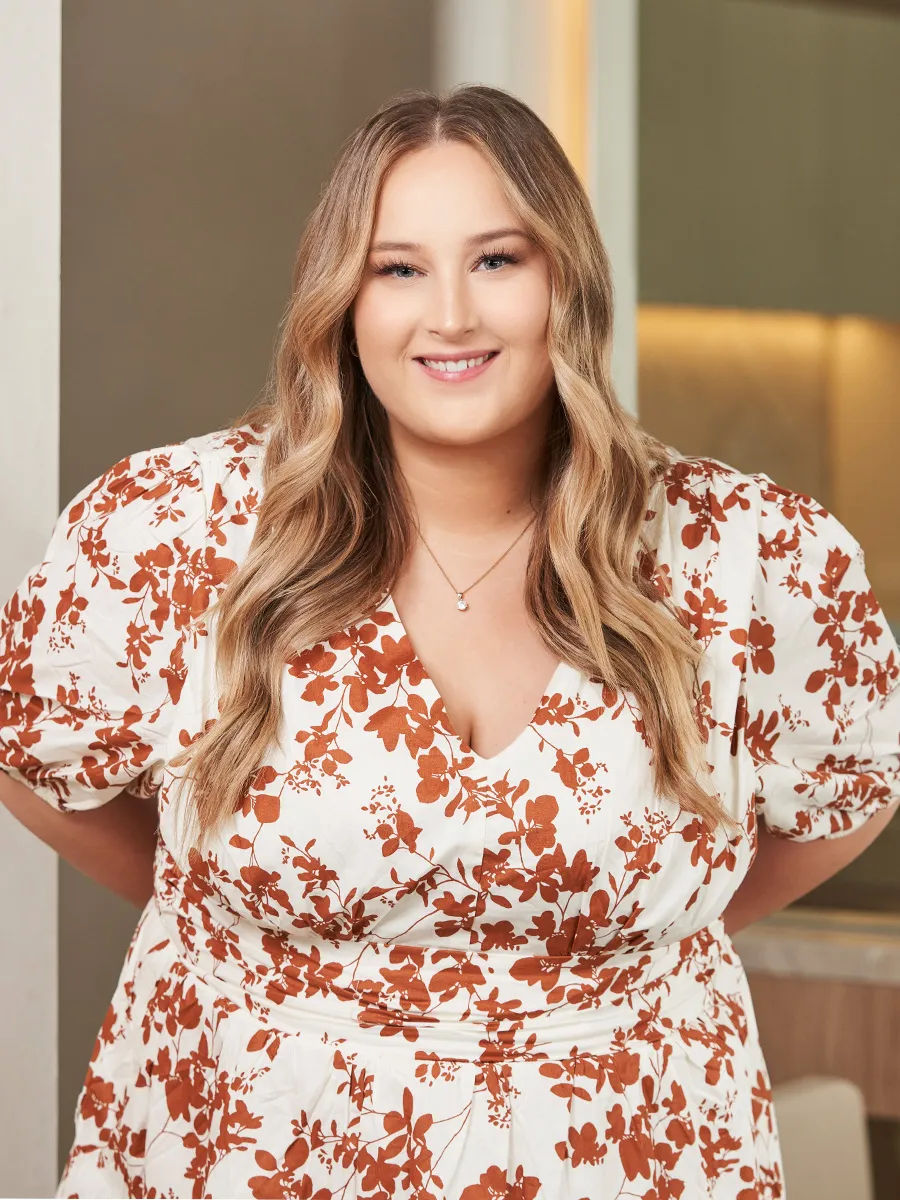 Smiling woman with long wavy blonde hair wearing a white dress with orange floral patterns and a pendant necklace.