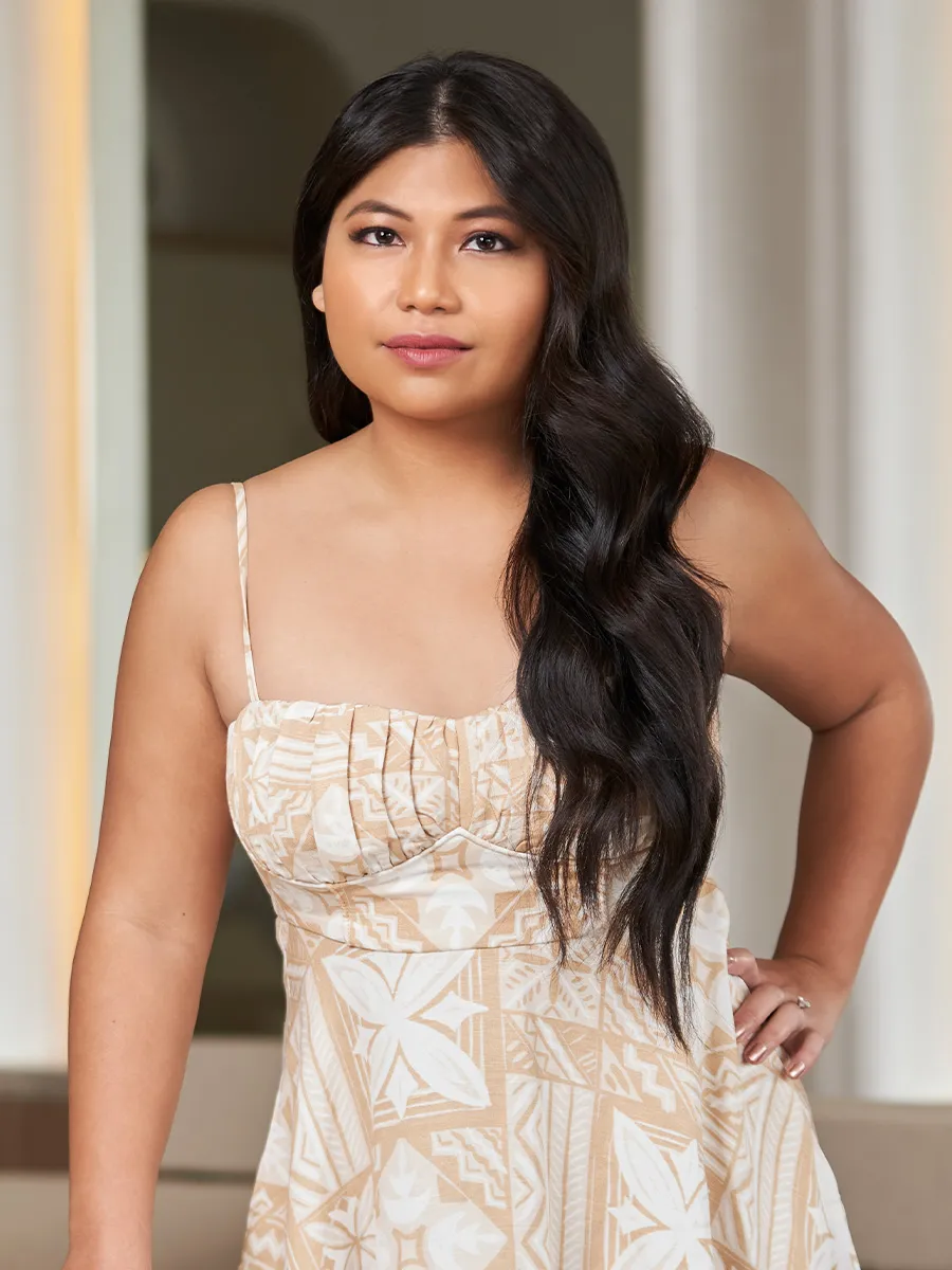 Woman with long dark wavy hair wearing a beige and white patterned sleeveless dress, standing indoors with one hand on her hip.