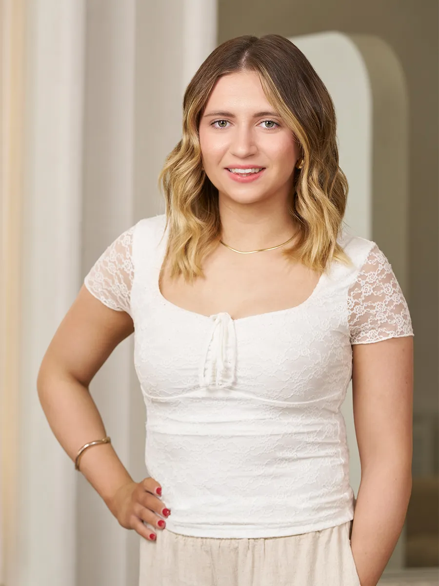 Smiling young woman with wavy blonde hair wearing a white lace top and beige pants, standing with hand on hip.