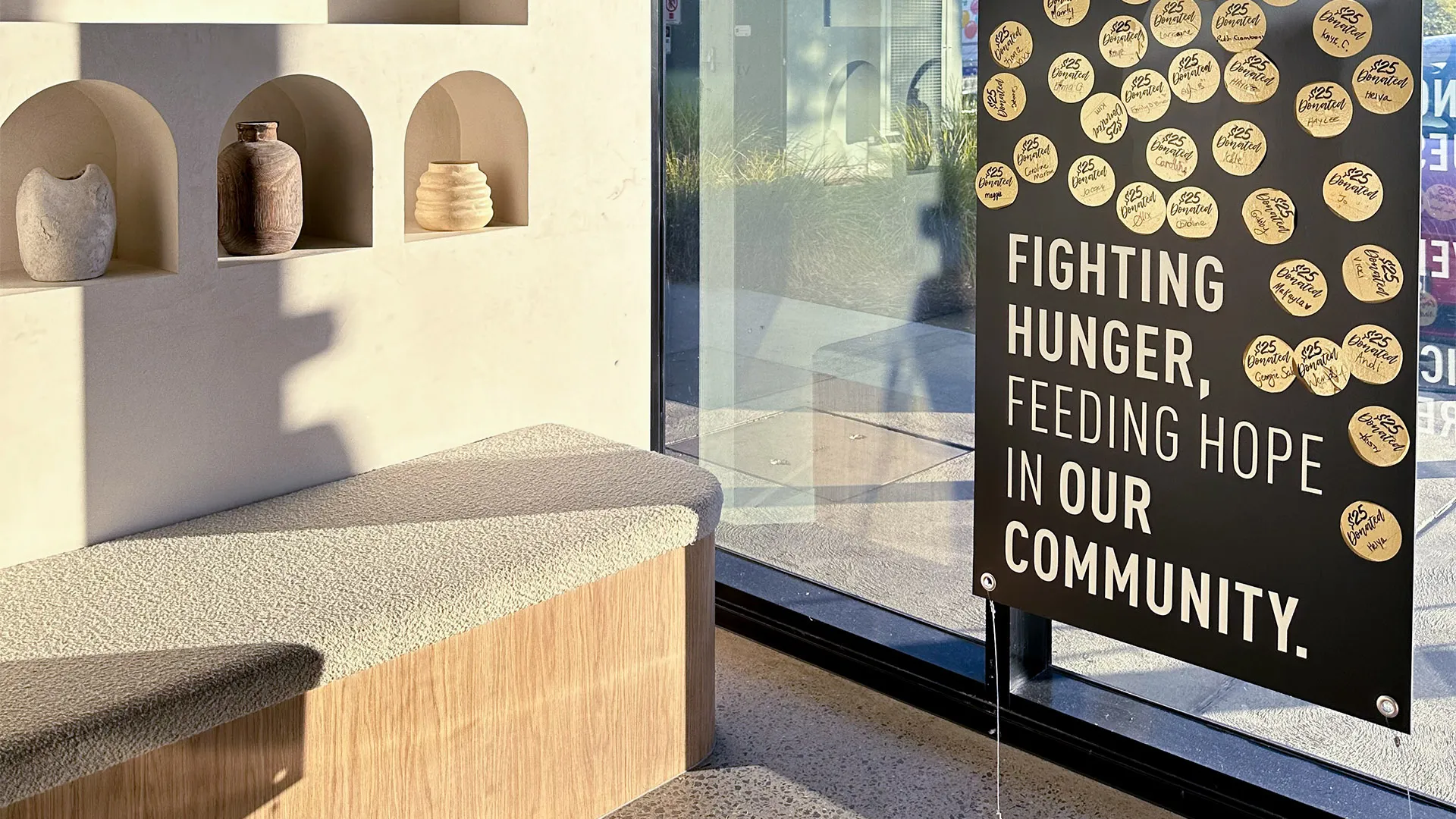 Indoor bench with textured cushion beside a wall with three decorative vases in arched niches and a window displaying a sign reading 'Fighting hunger, feeding hope in our community' with $25 donor stickers.
