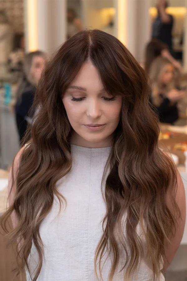 Woman with long, wavy brown hair and closed eyes wearing a light sleeveless top in an indoor setting.