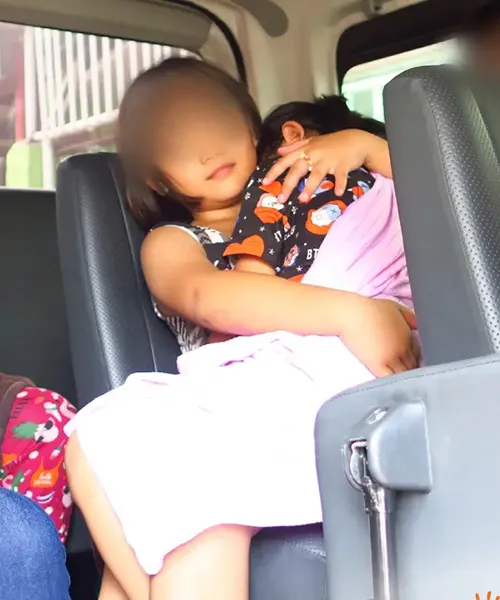 A young child sitting in a vehicle seat holding a baby wrapped in a pink blanket.