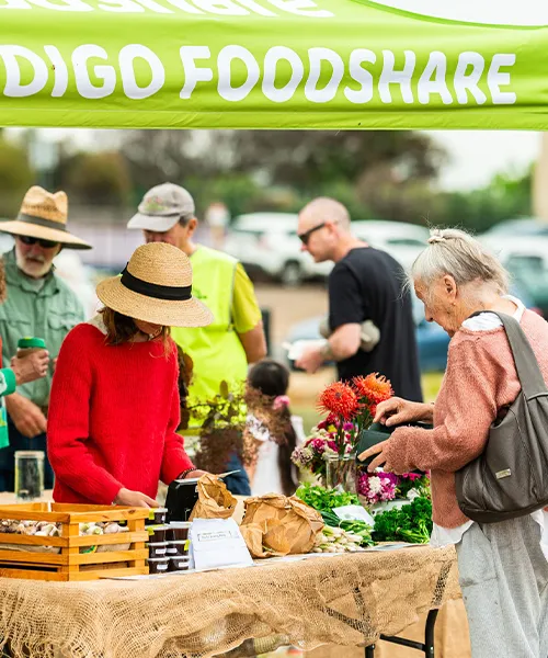 People interacting at a food share market stand with fresh produce and flowers under a green canopy labeled 'Foodshare'.