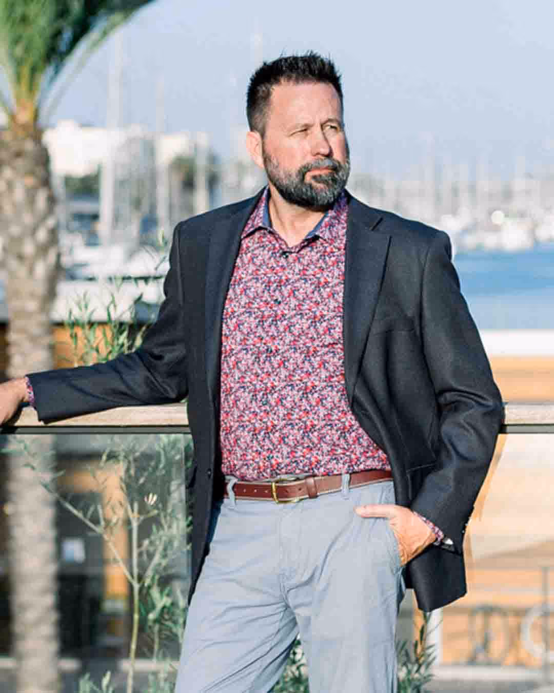 Man with beard wearing a floral shirt, black blazer, and light pants, standing outdoors by a railing with a marina in the background — Branden Lowder, CIAS real estate investor instructor at US Realty Training