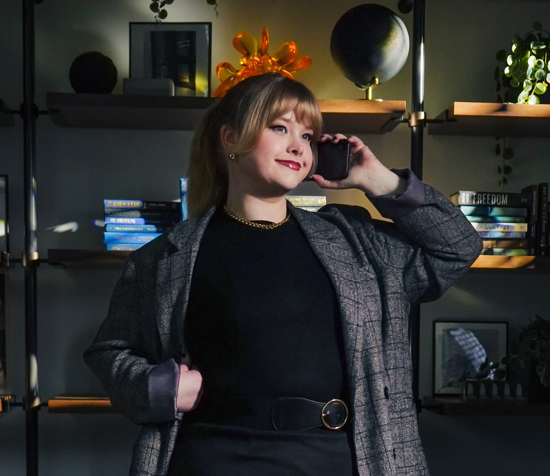 Young woman wearing a gray checked blazer and black belt, smiling while holding a phone to her ear in a cozy room with shelves and books.