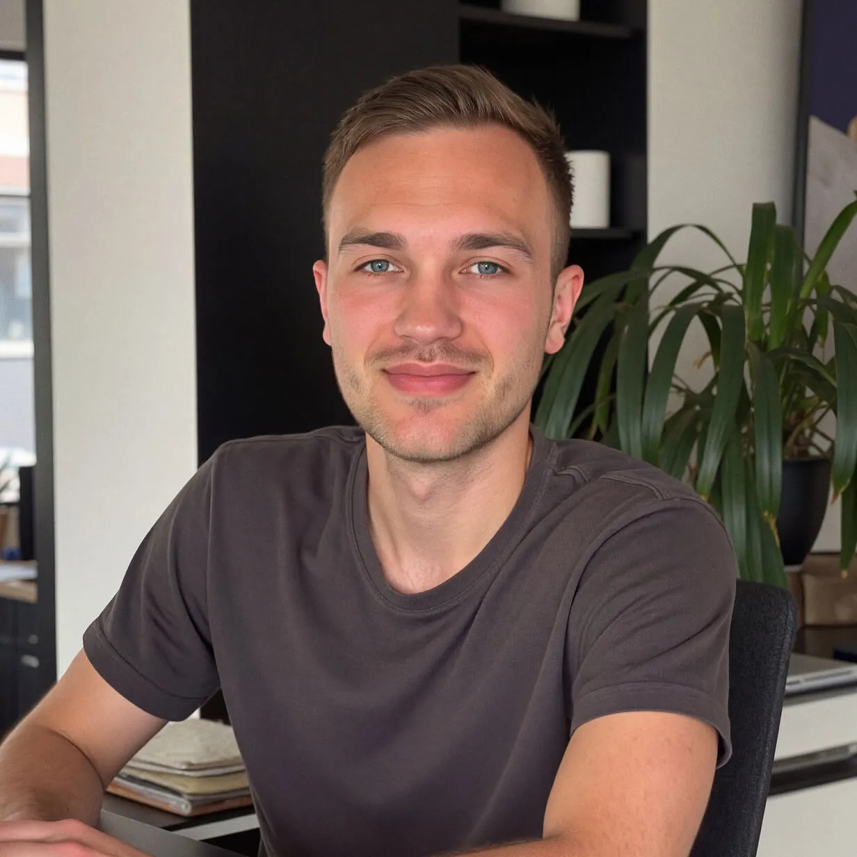 Roberto Dekker sitting at a desk indoors, smiling at the camera, wearing a dark T-shirt, with a plant and shelves in the background.