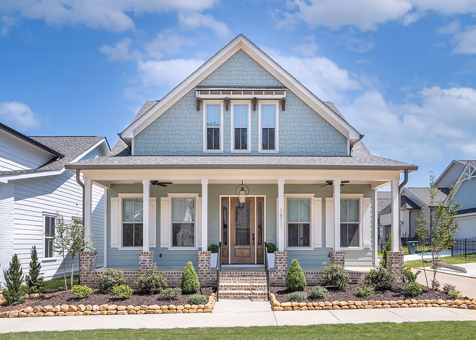 Blue two-story house with a front porch, brown door, brick steps, and landscaped garden beds bordered with stones.