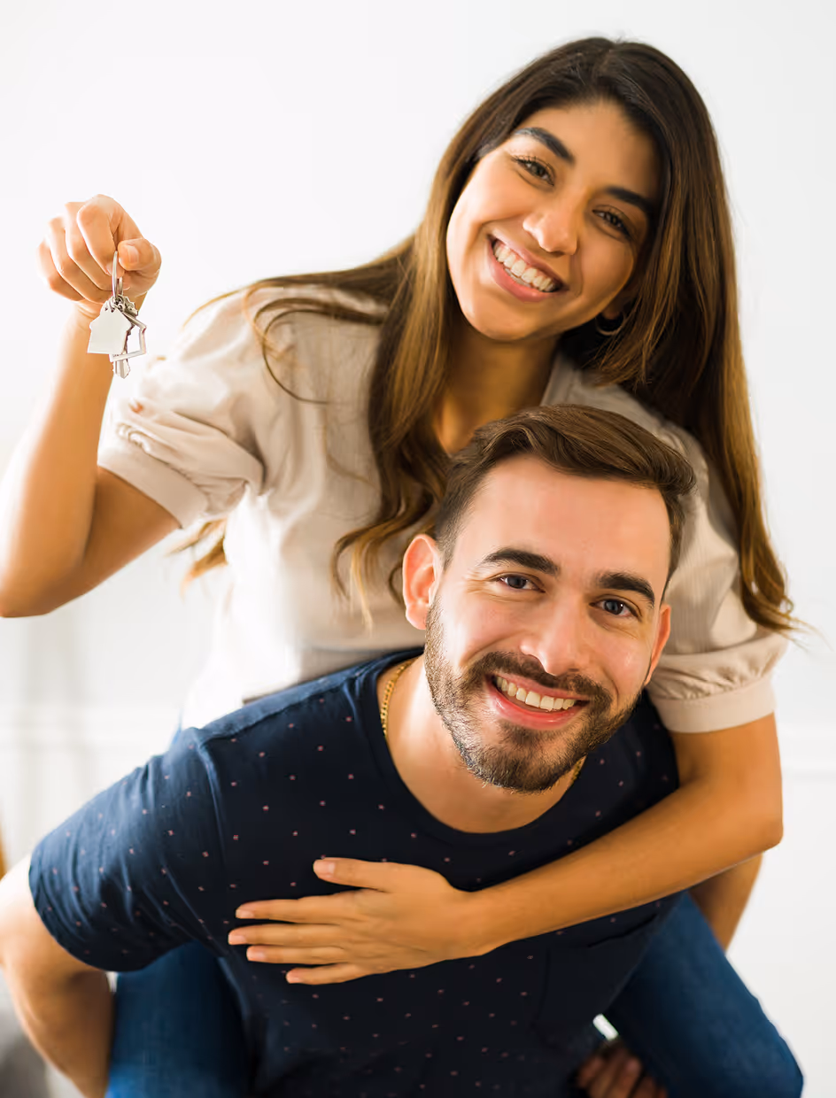 Smiling couple celebrating new home with woman holding house-shaped keys and man giving piggyback ride.